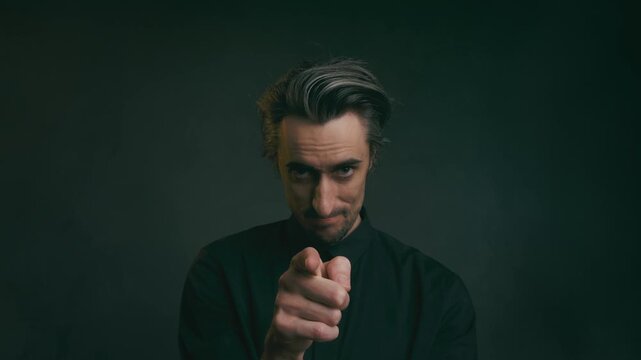 Confident young adult man pointing at camera in dark button-up shirt with blurred background and soft lighting, representing authority and motivation concepts.