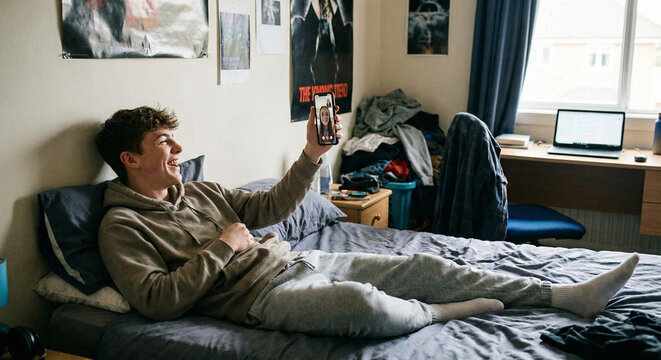 Happy teenage boy lying on bed video calling friend with smartphone in messy dorm room college student lifestyle.