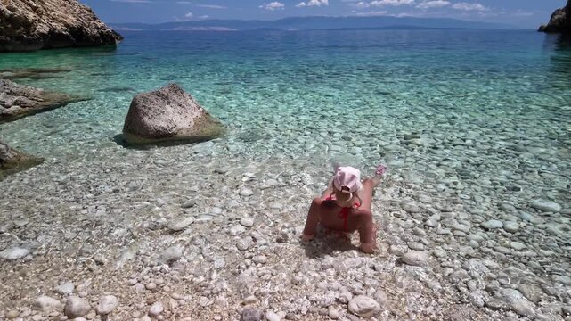 Woman sitting on pebble beach with feet immersed in serene Adriatic Sea. Enjoying a summer vacation in a hidden cove surrounded by natural rocky cliffs under bright blue sky in Cres Island of Croatia