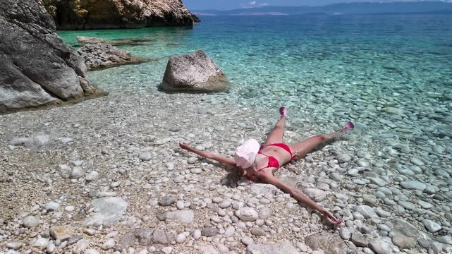 Woman sitting on pebble beach with feet immersed in serene Adriatic Sea. Enjoying a summer vacation in a hidden cove surrounded by natural rocky cliffs under bright blue sky in Cres Island of Croatia