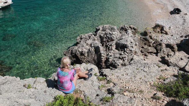Woman relaxing on a rocky coast, overlooking Sveti Ivan Beach with its crystal clear Adriatic Sea and moored sailboats, enjoying a peaceful summer day in Lubenice, Cres Island of Croatia
