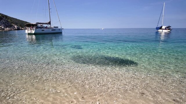 Hidden along rugged shores of Cres Island in Croatia, Sveti Ivan Beach features transparent turquoise sea, lush forested hills, and handful of boats resting in calm waters beside the remote pebble bay