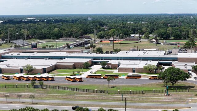 PCHS lettering marks perimeter fence outside Plant City Florida high school, row of parked yellow buses lines lot as cars pass road beside baseball and football fields. Aerial drone wide shot