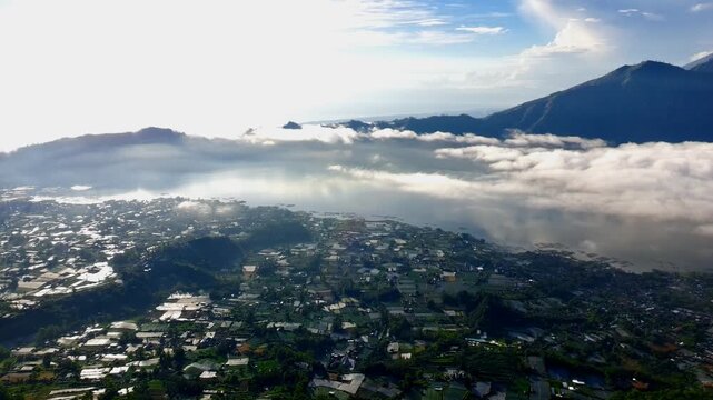 Aerial view of Danau Batur surrounded by lush mountains and low-hanging clouds in the volcanic landscape of Kintamani, Bali, Indonesia.