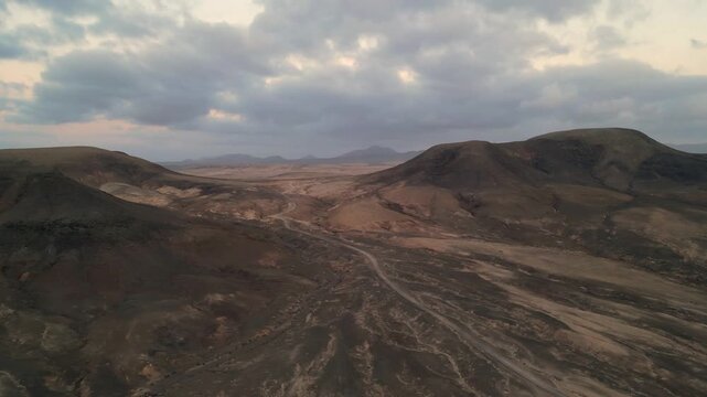 Aerial view of the arid volcanic landscape with rolling hills and a dirt road under a cloudy sky in El Cotillo, Canary Islands, Spain.