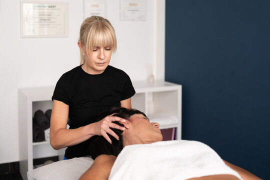 Female therapist performing manual neck therapy on adult patient in calm treatment room with focused expression, white sheet, soft directional lighting, and clinical wellness setting