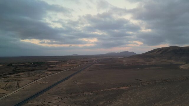 Aerial view of the volcanic landscape near El Cotillo with a long road stretching through the arid terrain under a dramatic cloudy sky in El Cotillo, Canary Islands, Spain.