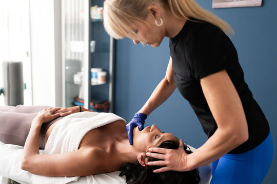 Therapist performing gentle facial massage on relaxed woman lying on treatment table in modern wellness clinic with natural light, blue walls, towel covering chest