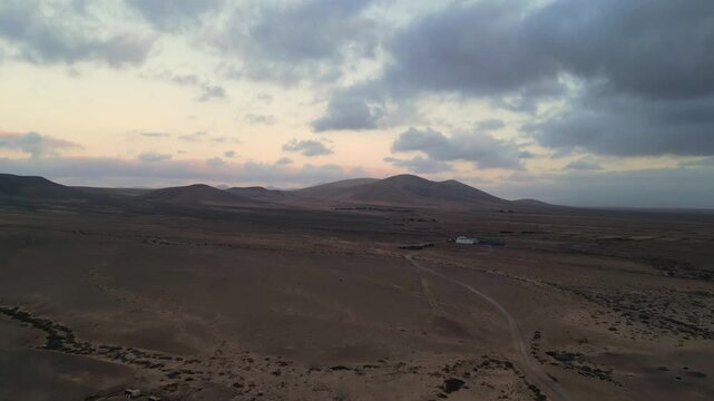 Aerial view of the arid volcanic desert landscape with rolling hills and a winding dirt road under a cloudy sunset sky in El Cotillo, Canary Islands, Spain.