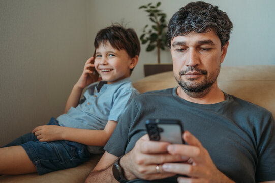 Father uses smartphone while son plays with cardboard phone at home
