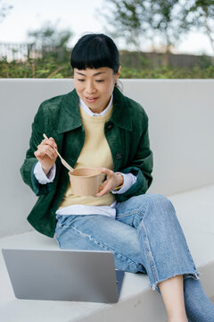 Smiling woman sitting outside eating lunch and using laptop