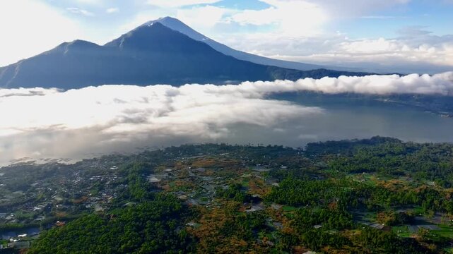 Aerial view of Danau Batur lake surrounded by lush green forests and volcanic mountains under a bright sky in Kintamani, Bali, Indonesia.