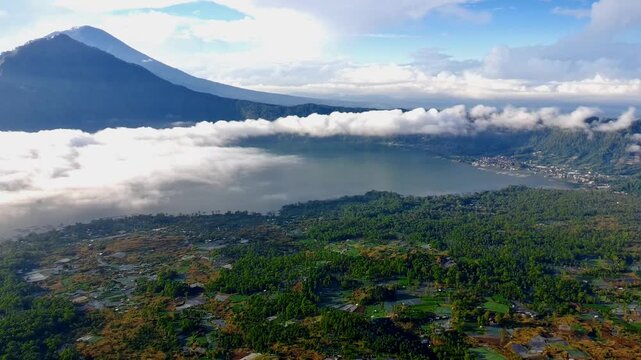 Aerial view of Danau Batur surrounded by lush green mountains and low-hanging clouds in Kintamani, Bali, Indonesia.