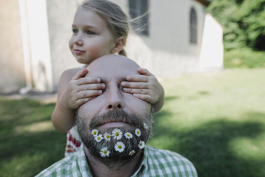 Little girl's hands covering eyes of mature man with daisies in his beard