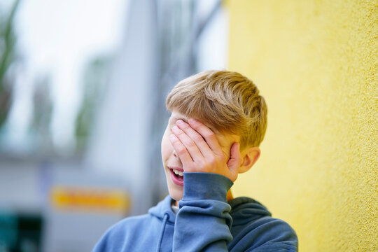 Blond boy covering face with hand in front of yellow wall