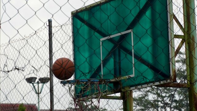 Basketball at the rim of an old outdoor hoop behind a chain link fence, showing weathered sports equipment at an urban court.