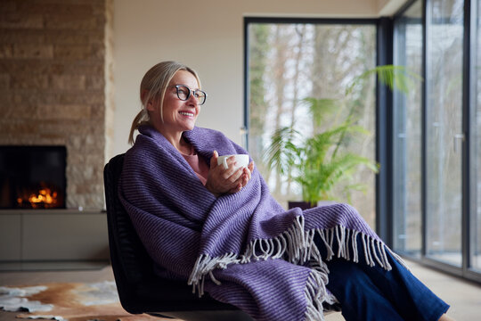 Happy blond woman wrapped in blanket holding coffee cup sitting at home