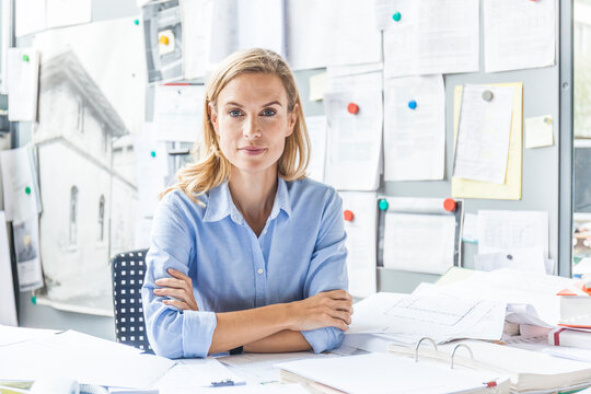 Portrait of confident woman sitting at desk in office surrounded by paperwork