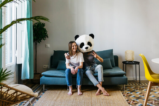 Woman wearing panda mask gesturing peace sign while sitting with girlfriend at home