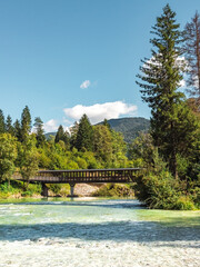 Fototapeta premium Wooden pedestrian bridge crossing the turquoise waters of the Sava Bohinjka River surrounded by lush forest and alpine scenery