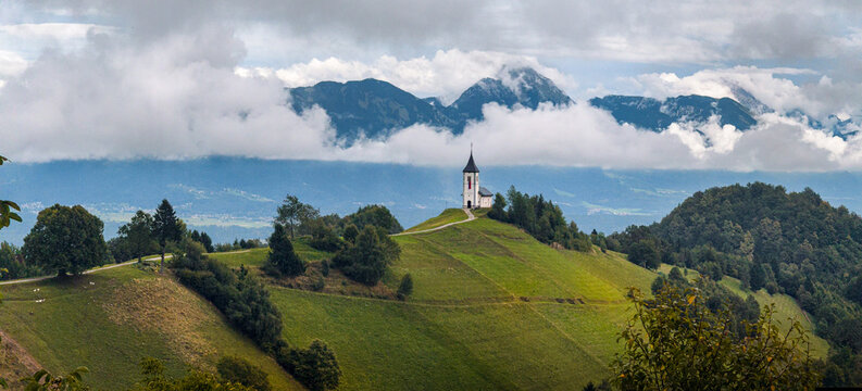 Scenic Church of Saint Primoz standing on Jamnik Hill above rolling green hills with the Julian Alps rising through dramatic clouds