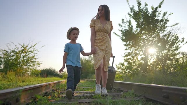 Mother and son walking on railway track with suitcase. Family holding hands on railroad starts summer travel. Woman and boy go on vacation journey. Parent and child walk forward on sunny train track.