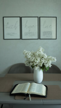Fresh white lilac bouquet in vase beside opened holy bible scripture on wooden table in bright home dining space. JPG