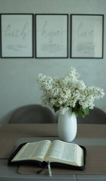 Minimalist living room display featuring white lilac flowers in vase and opened holy bible scripture on dark table. JPG