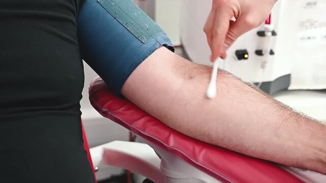 Footage of a hand cleaning the inner elbow of a donor with a cotton swab above a tourniquet, before a blood donation at a medical clinic.