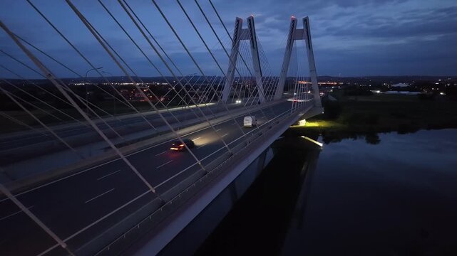 Nighttime view of a modern cable-stayed bridge with vehicles passing over the roadway and reflections visible in the calm water below. Car riding at dark highway road. Auto driving at night route