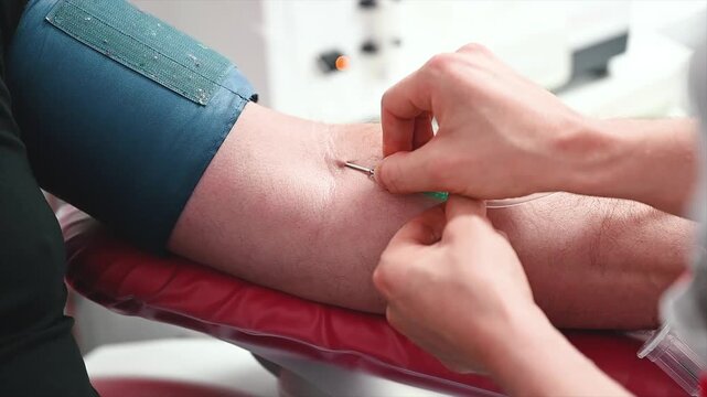 Footage of gloved hands inserting the donation needle into the vein of a donor arm during a blood donation at a clinic, the start of the procedure.