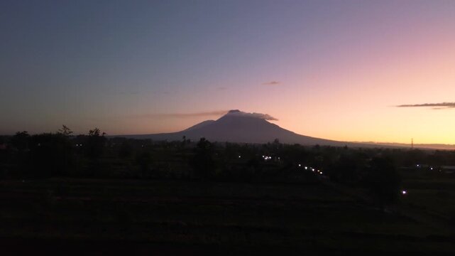 A stunning panoramic view of Mount Merapi from a distance, set against a dark pre-dawn sky. Aerial footage capturing the beauty of Mount Merapi while the sky is still dark.