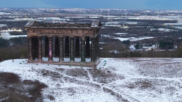 Telephoto drone orbit shot of the snow-covered Penshaw Monument in Sunderland, with the Nissan car factory and wind turbines visible in the compressed background.