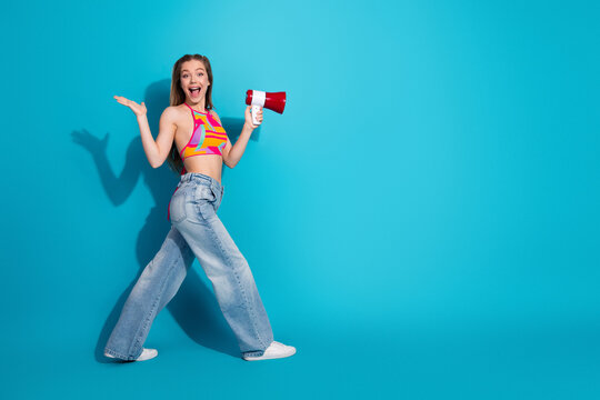 Young woman joyfully posing with a megaphone against a vibrant blue background in stylish and trendy casual attire