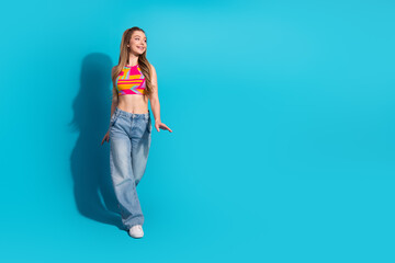 Obraz premium Young cheerful female in colorful top and jeans standing against vibrant blue background exuding happiness and style