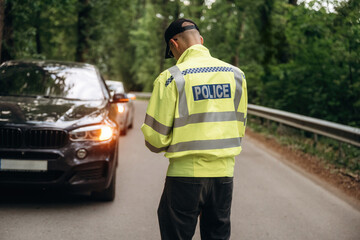 Obraz premium Police officer in reflective yellow jacket stands on roadside checking vehicle details with cars parked in a forested area, trees lining the road