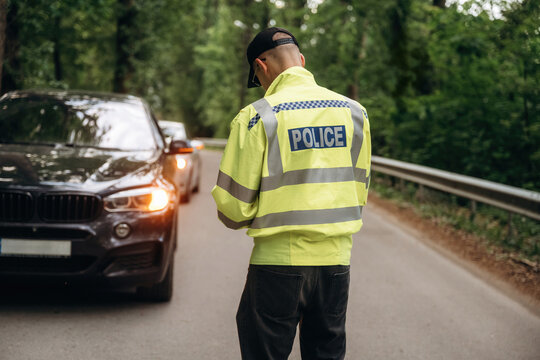 Police officer in reflective yellow jacket stands on roadside checking vehicle details with cars parked in a forested area, trees lining the road