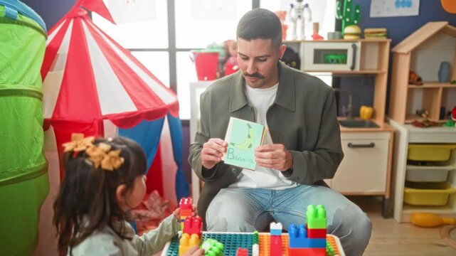 Man reading a flashcard and pointing finger to child in building playroom with colorful blocks and tent; learning play joy.