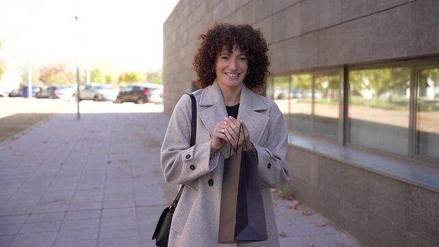 A stylish woman with curly hair in an urban setting holds a shopping bag of delightful items