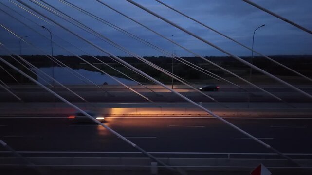 Cars driving at night across a modern bridge with cables, showcasing a scenic view of the river and landscape under a twilight sky. Vehicles riding at dark highway road. Auto driving at night route