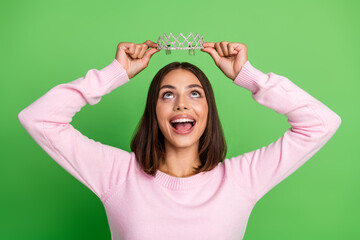 Young stylish woman in pink sweater crowns herself with a tiara smiling against green studio background © deagreez