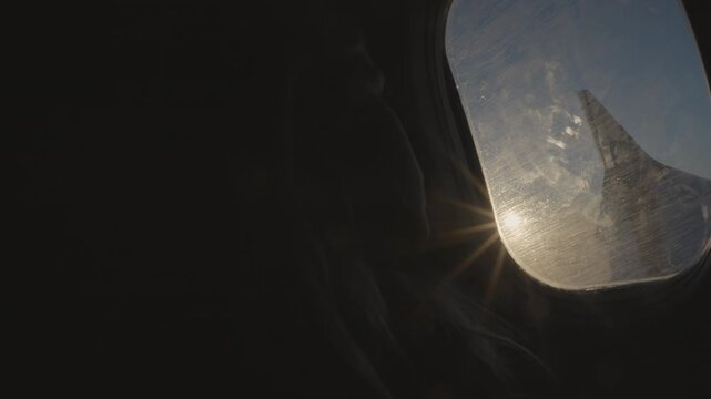 Silhouette of a passenger looking out of an airplane window during flight, showing a bright sun flare and the wing against a blue sky