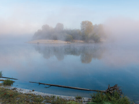 A misty morning landscape captures a small, tree-covered island gently reflected in the calm, foggy waters of a serene river, with a fallen log resting onshore