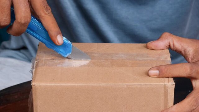 Close-up of an Asian man's hands opening a sealed cardboard box with a utility knife during a home package unboxing. Parcel delivery, online shopping, and package opening.
