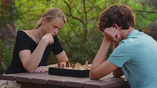 A mother and her fifteen year old teenage son play chess together at a wooden table in a park, focusing on their strategy and next move