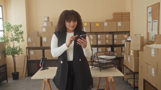 Woman tapping smartphone with finger at parcel packing desk amid stacked cardboard boxes in building; small business confidence.