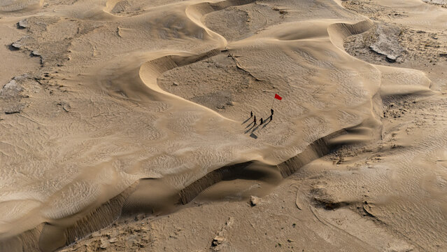 Aerial view of a group of people standing on sand dunes in the desert with a red flag, casting long shadows in the sunlight, Al Neser, Dhi Qar Governorate, Iraq.