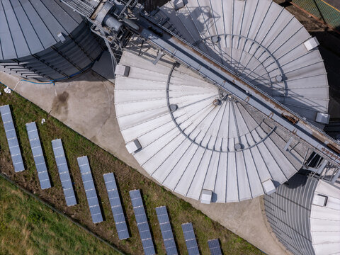 Aerial view of industrial grain silos with metallic roofs and rows of solar panels on the ground in Grudziadz, Kuyavian-Pomeranian Voivodeship, Poland.