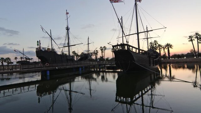 Cinematic wide shot of the historical replica ships. Pinta, Ni&ntilde;a and Santa Mar&iacute;a docked at Muelle de las Carabelas during a serene sunset. Located in La Rabida, Palos de la Frontera, Huelva, Spain. 