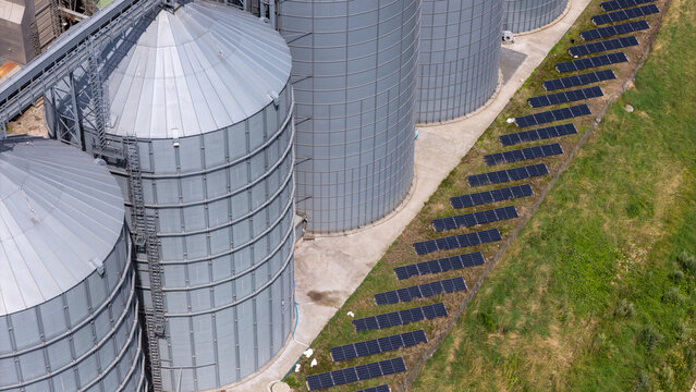 Aerial view of large industrial grain silos with a row of solar panels installed on the grassy ground in Grudziadz, Kuyavian-Pomeranian Voivodeship, Poland.